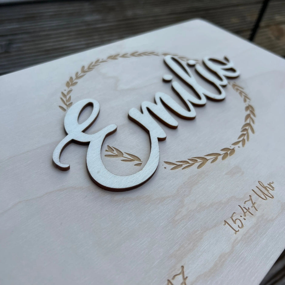 Personalisierte Holztafel mit dem Namen Emilio im 3D-Schriftzug, umrahmt von einem Blätterkranz
